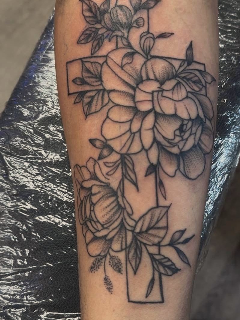 Black-and-grey cross intertwined with peony flowers on a forearm.